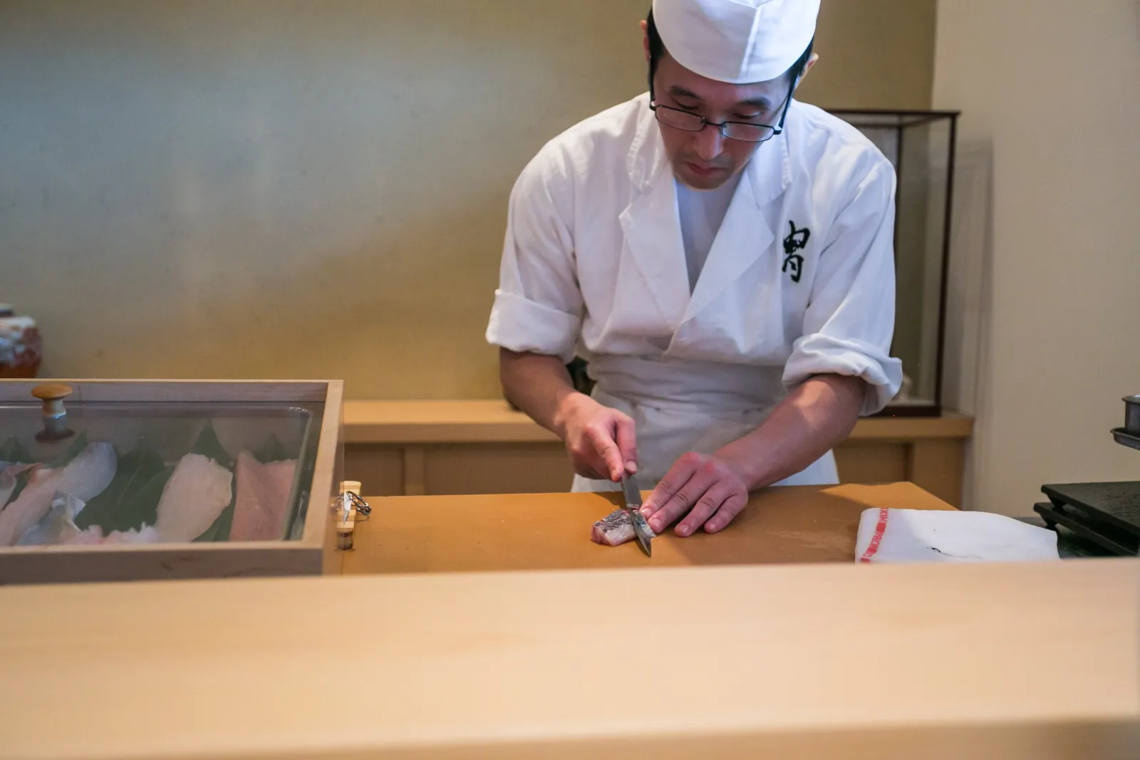 A sushi chef slicing fish at a pale hinoki counter